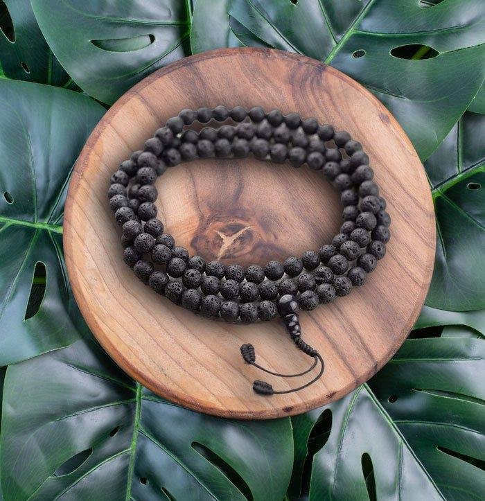 A dark prayer bead mala coiled on a wooden plate over monstera leaves.