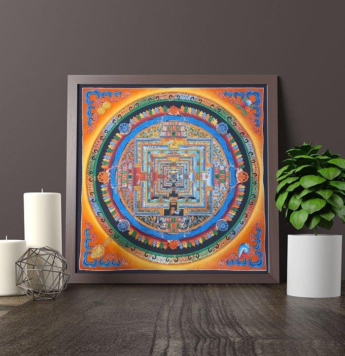 A framed Kalachakra mandala leaning on a dark wooden sideboard with candles and a plant.
