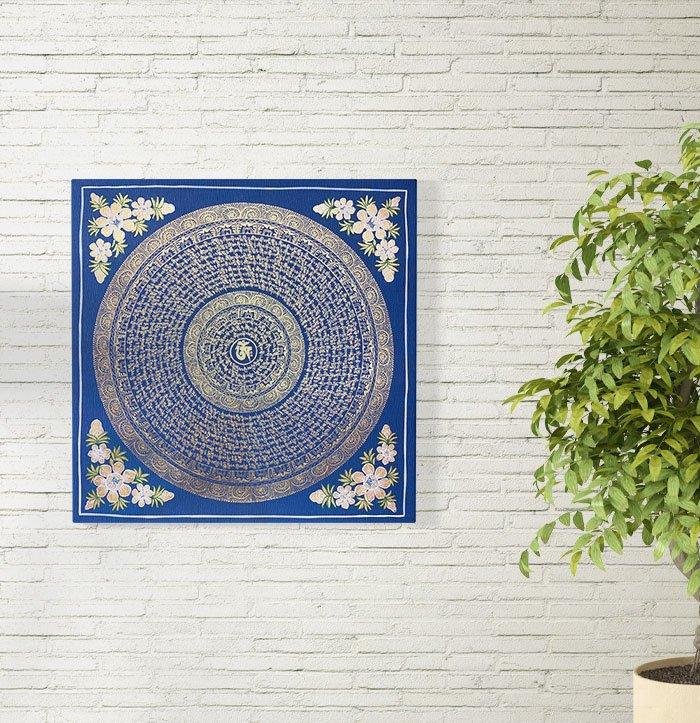 A blue and silver mantra mandala hanging on a white brick wall.