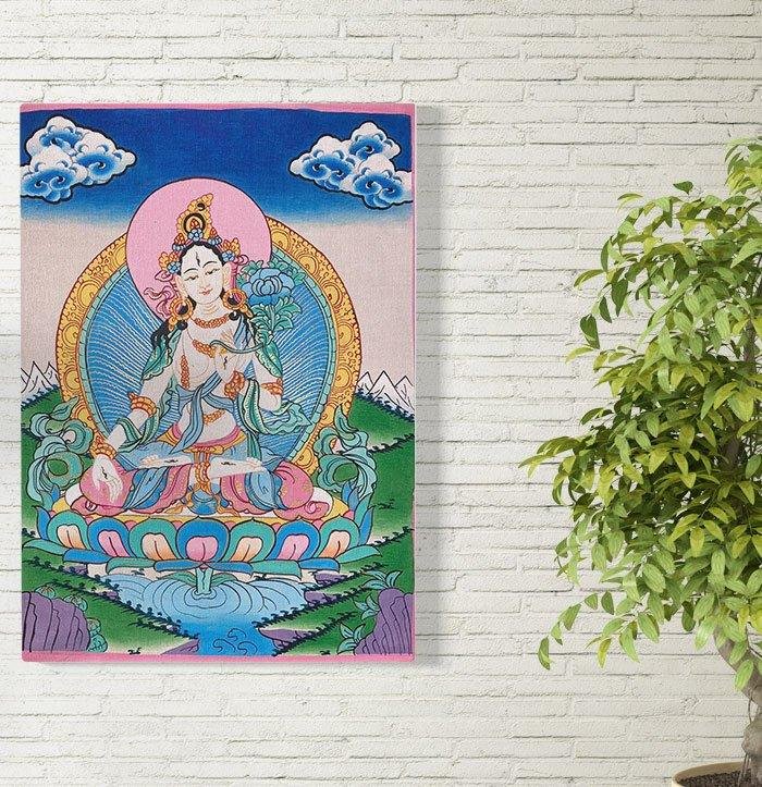 A colourful Green Tara thangka hanging on a white brick wall beside a green plant.